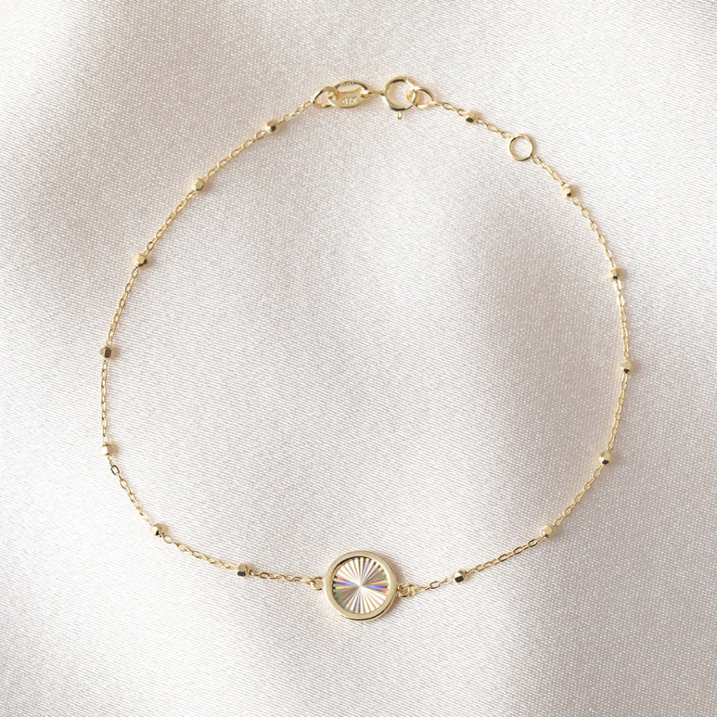 A delicate gold chain bracelet with small bead accents and a central round disc, displayed on a soft white fabric background.