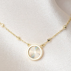 A delicate gold chain necklace displayed on a satin. The necklace features a round gold pendant with a sunburst pattern.