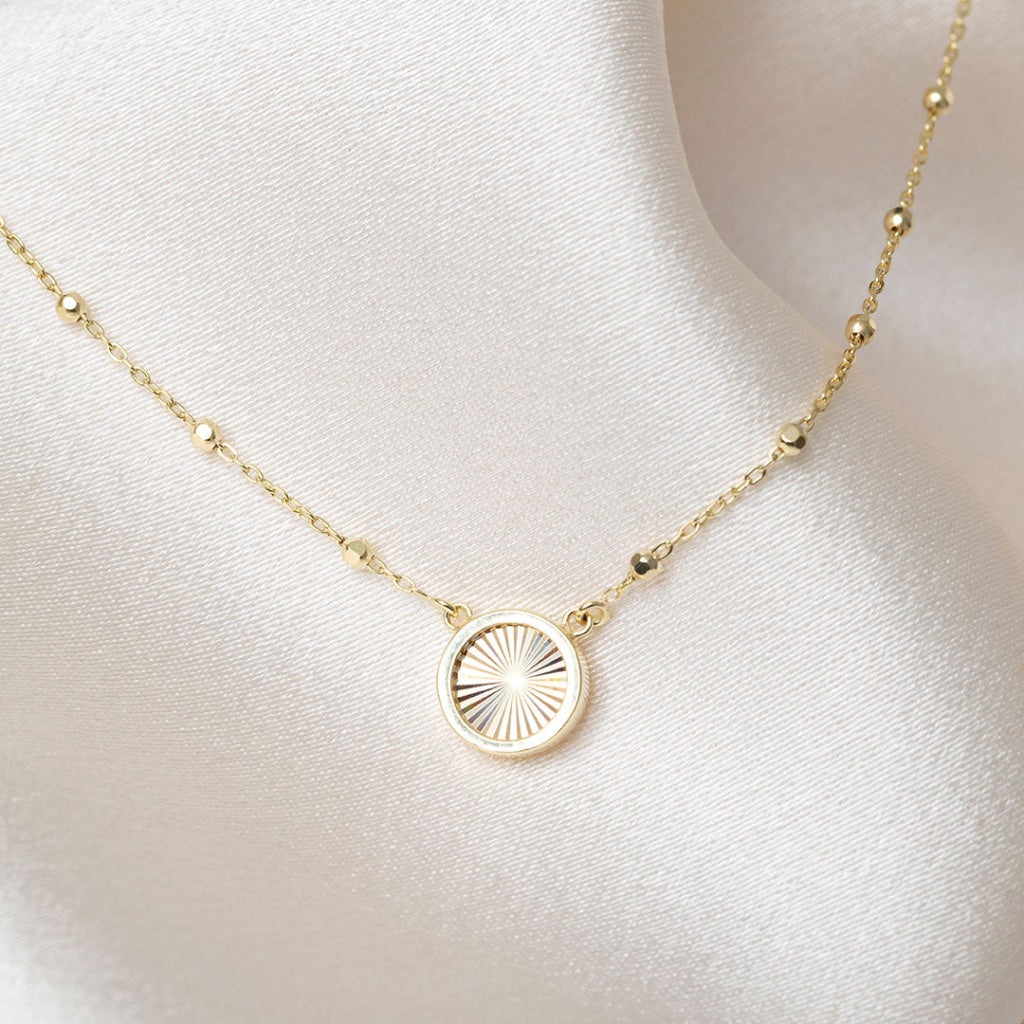 A delicate gold chain necklace displayed on a satin. The necklace features a round gold pendant with a sunburst pattern.