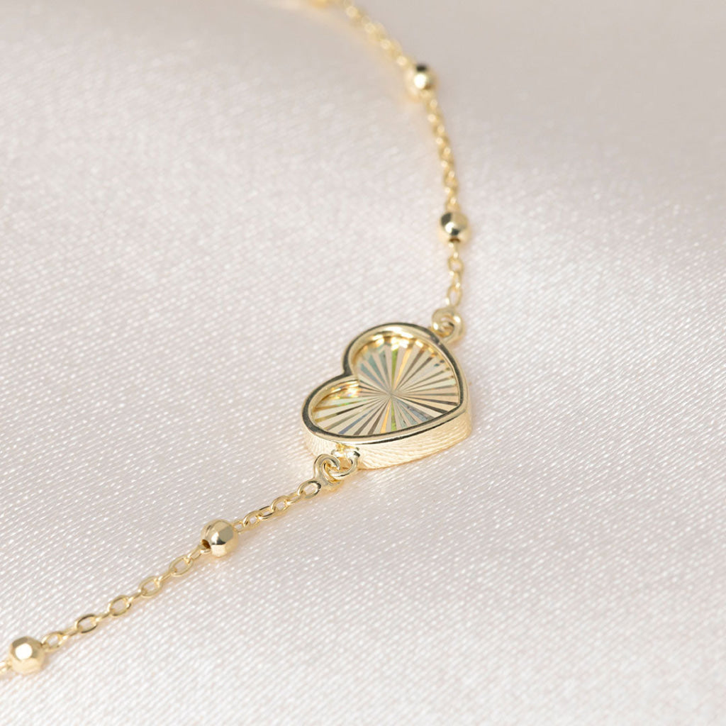 A delicate gold chain bracelet with a small heart-shaped pendant featuring a sunburst pattern, displayed on a soft, light-colored fabric background.