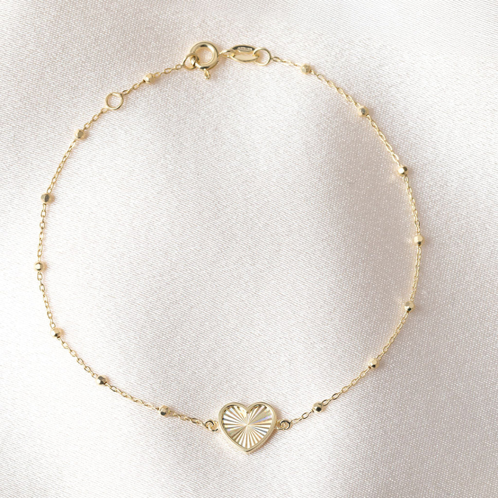 A delicate gold chain bracelet with a small heart-shaped pendant featuring a sunburst pattern, displayed on a soft, light-colored fabric background.