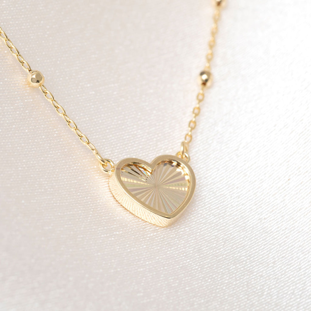 A delicate gold chain necklace with a small heart-shaped pendant featuring a sunburst pattern, displayed on a textured fabric background.
