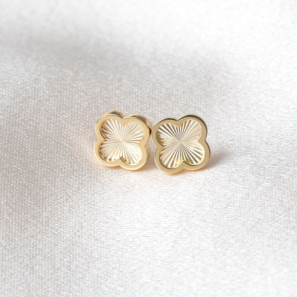 A pair of gold clover shaped stud earrings with a sunburst pattern on a textured white fabric background.