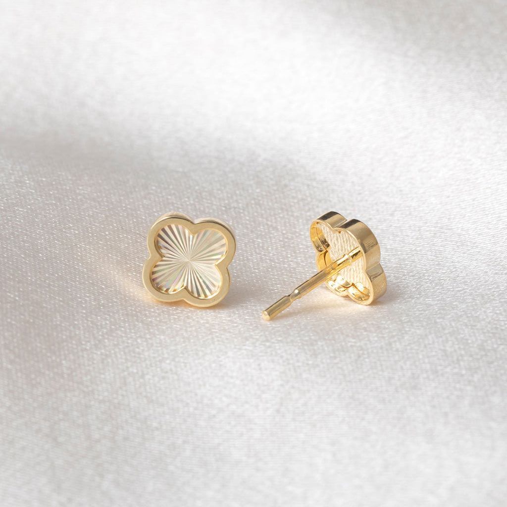 A pair of gold clover shaped stud earrings with a sunburst pattern on a textured white fabric background.