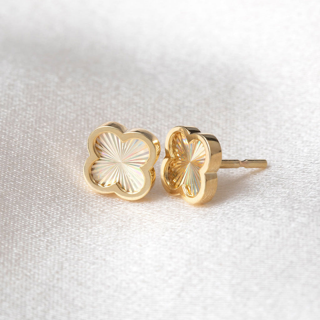 A pair of gold clover shaped stud earrings with a sunburst pattern on a textured white fabric background.