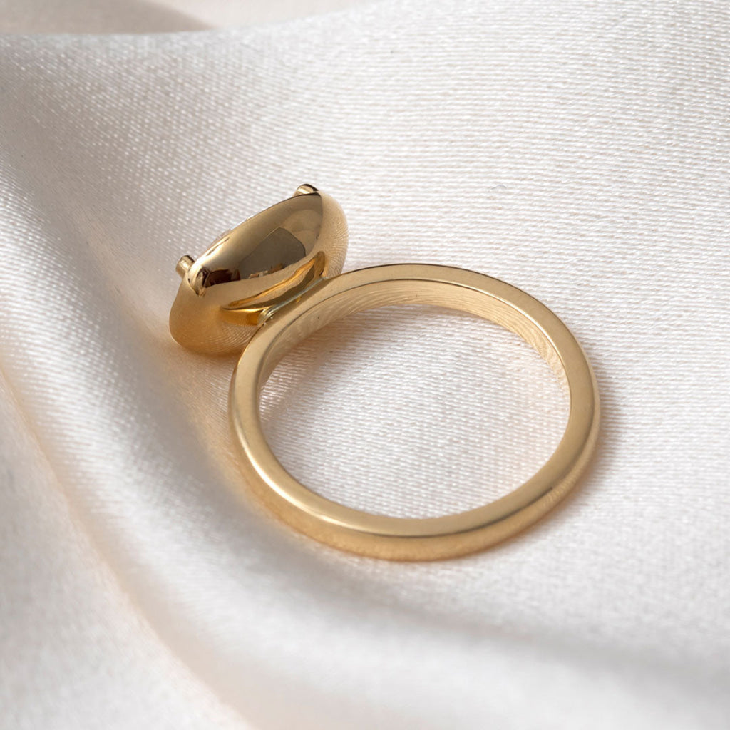 A gold ring with a polished, rounded dome-shaped top, resting on draped white fabric.