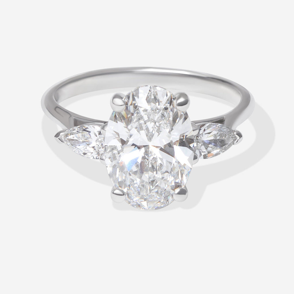 An oval diamond engagement ring with a platinum band featuring small pear-cut side diamonds, shown on a light grey background.