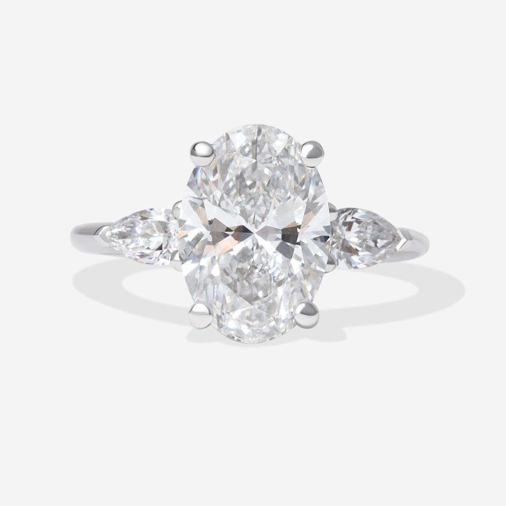 An oval diamond engagement ring with a platinum band featuring small pear-cut side diamonds, shown on a light grey background.