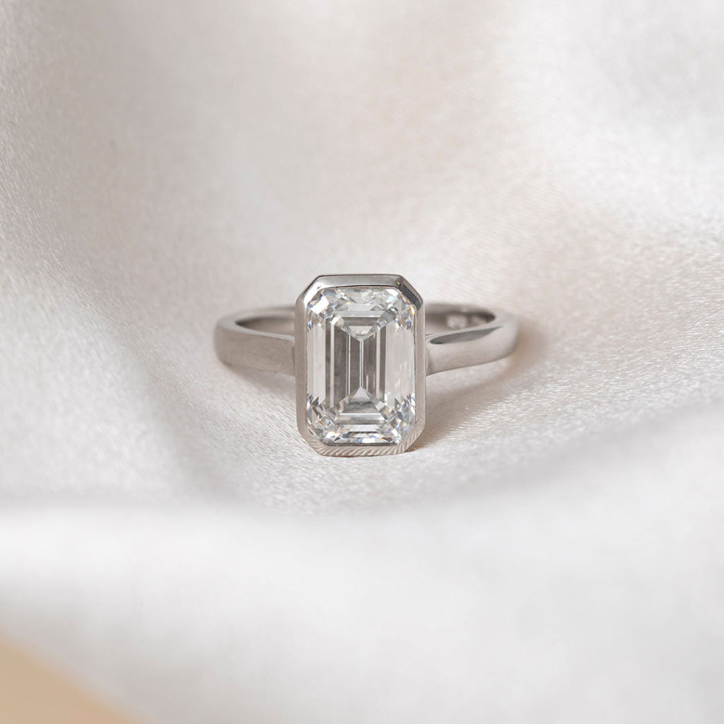An emerald-cut diamond ring in a platinum band, resting on a soft, textured white fabric background.