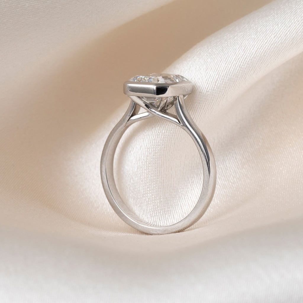 An emerald-cut diamond ring in a platinum band, resting on a soft, textured white fabric background.