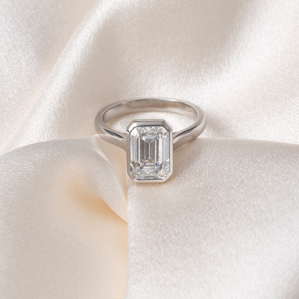 An emerald-cut diamond ring in a platinum band, resting on a soft, textured white fabric background.