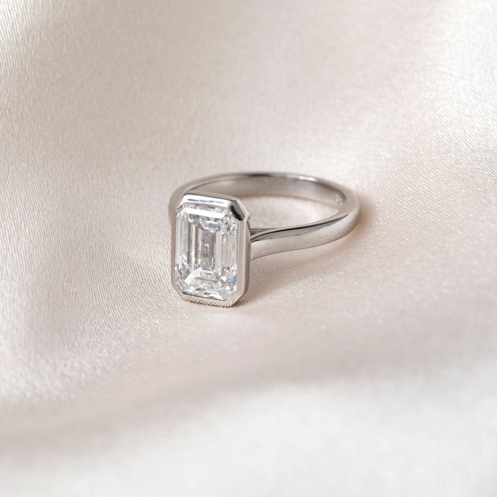 An emerald-cut diamond ring in a platinum band, resting on a soft, textured white fabric background.