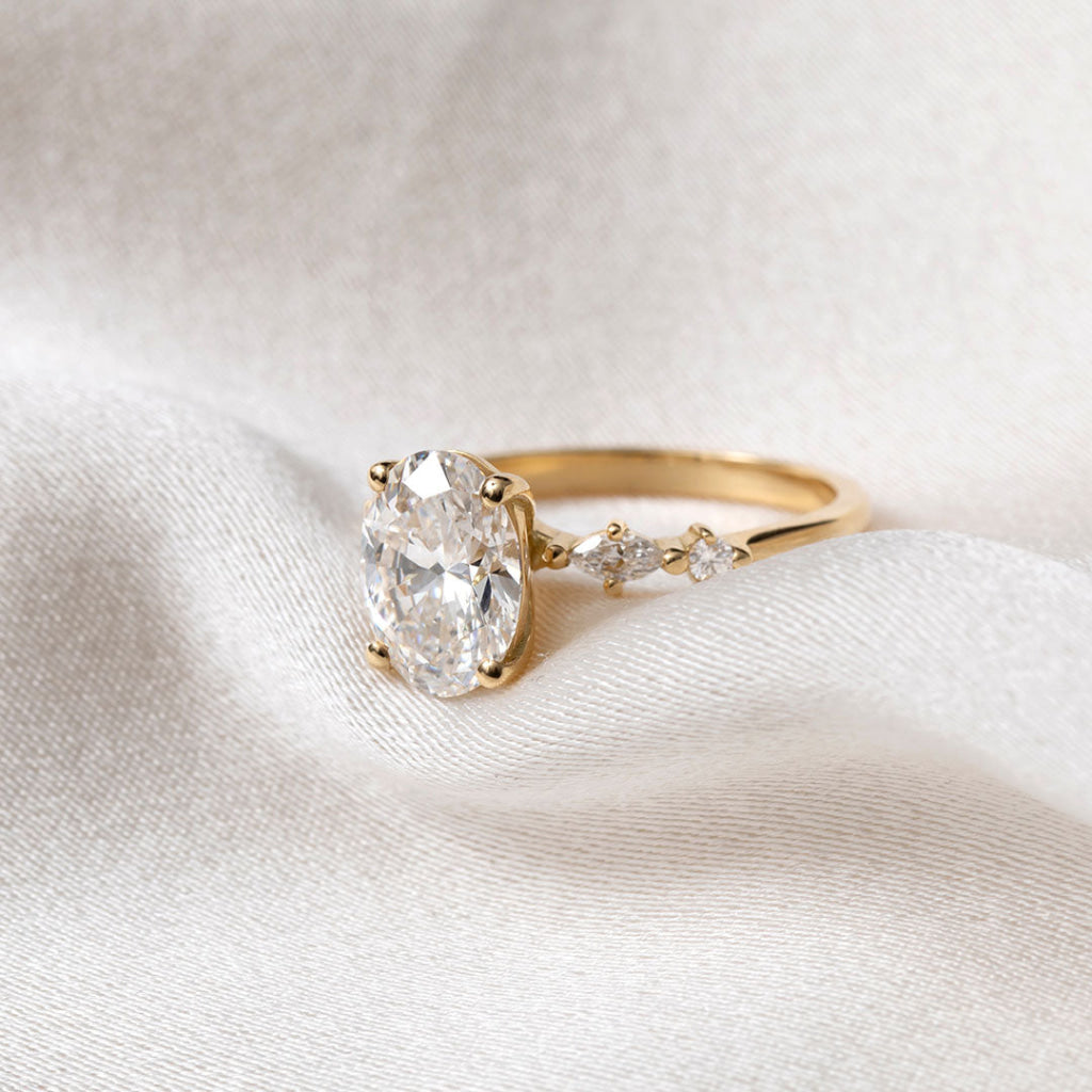 A yellow gold solitaire engagement ring with a round brilliant-cut center stone and marquise-cut side stones, resting on draped cream-colored satin fabric.