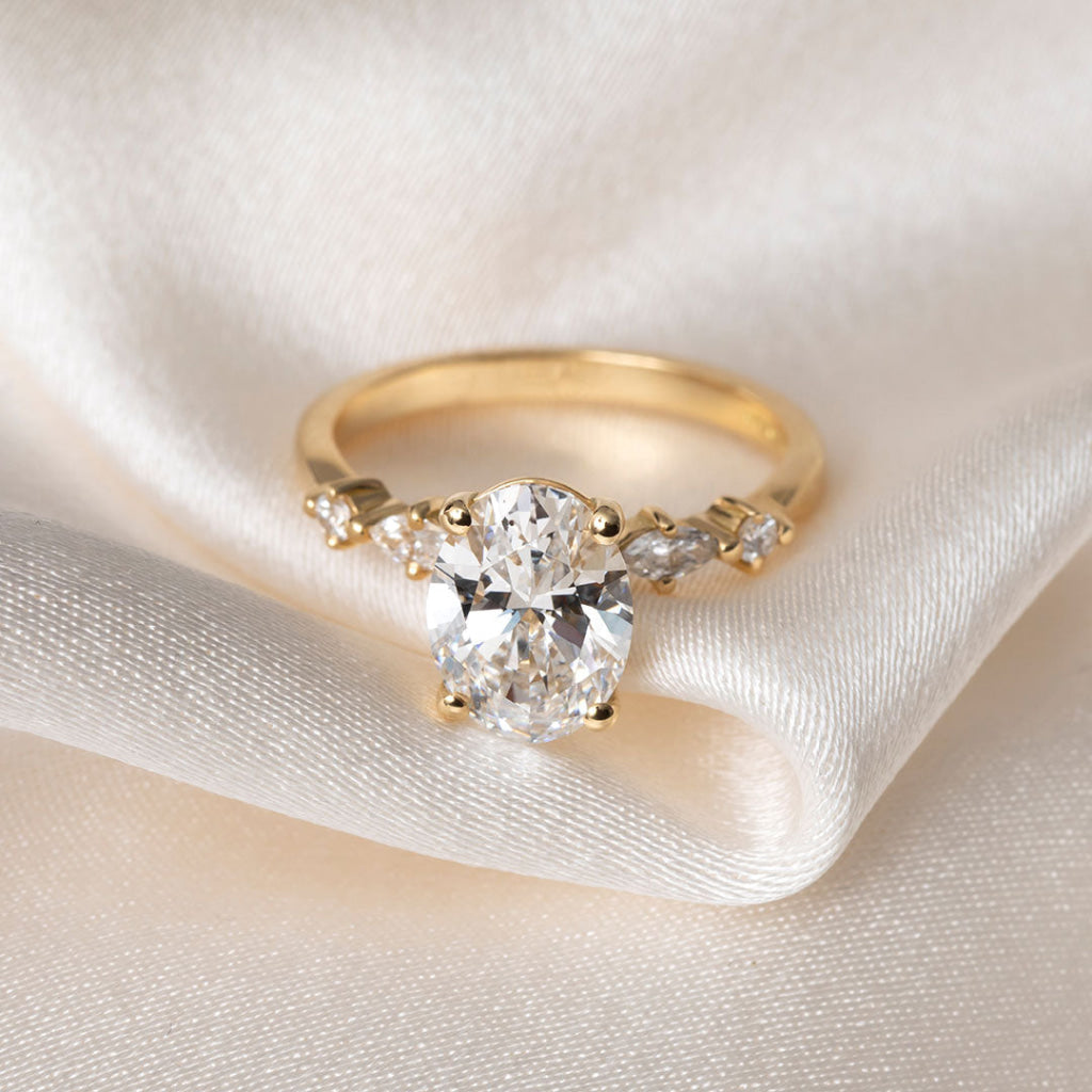 A yellow gold solitaire engagement ring with a round brilliant-cut center stone and marquise-cut side stones, resting on draped cream-colored satin fabric.