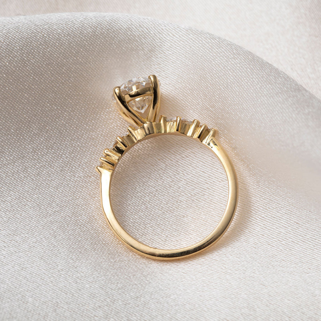 A yellow gold solitaire engagement ring with a round brilliant-cut center stone and marquise-cut side stones, resting on draped cream-colored satin fabric.