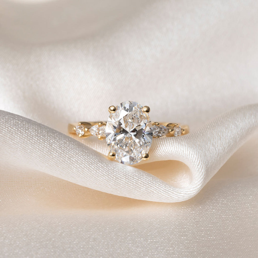 A yellow gold solitaire engagement ring with a round brilliant-cut center stone and marquise-cut side stones, resting on draped cream-colored satin fabric.