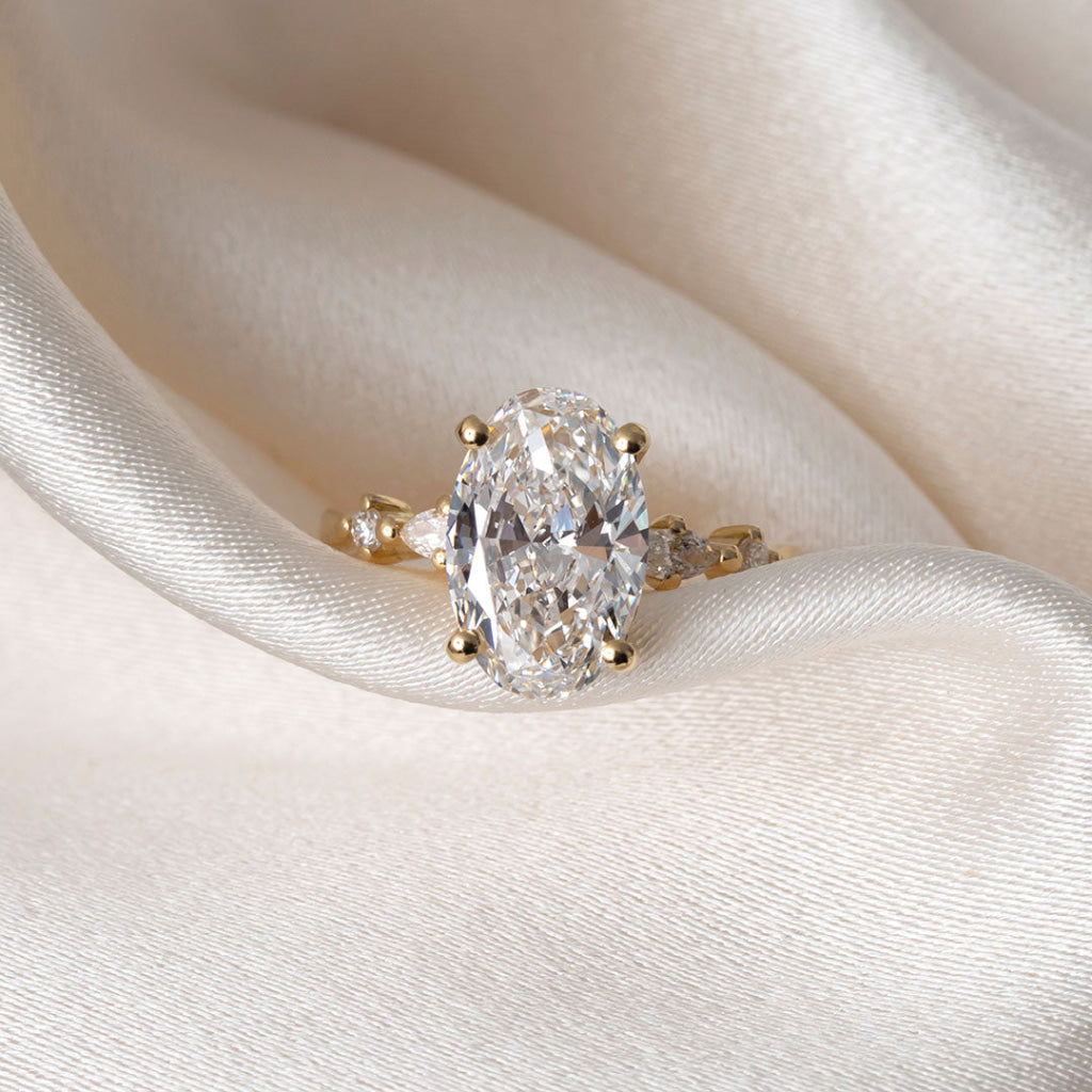 An oval diamond engagement ring with a yellow gold band, resting on draped cream-colored satin fabric.