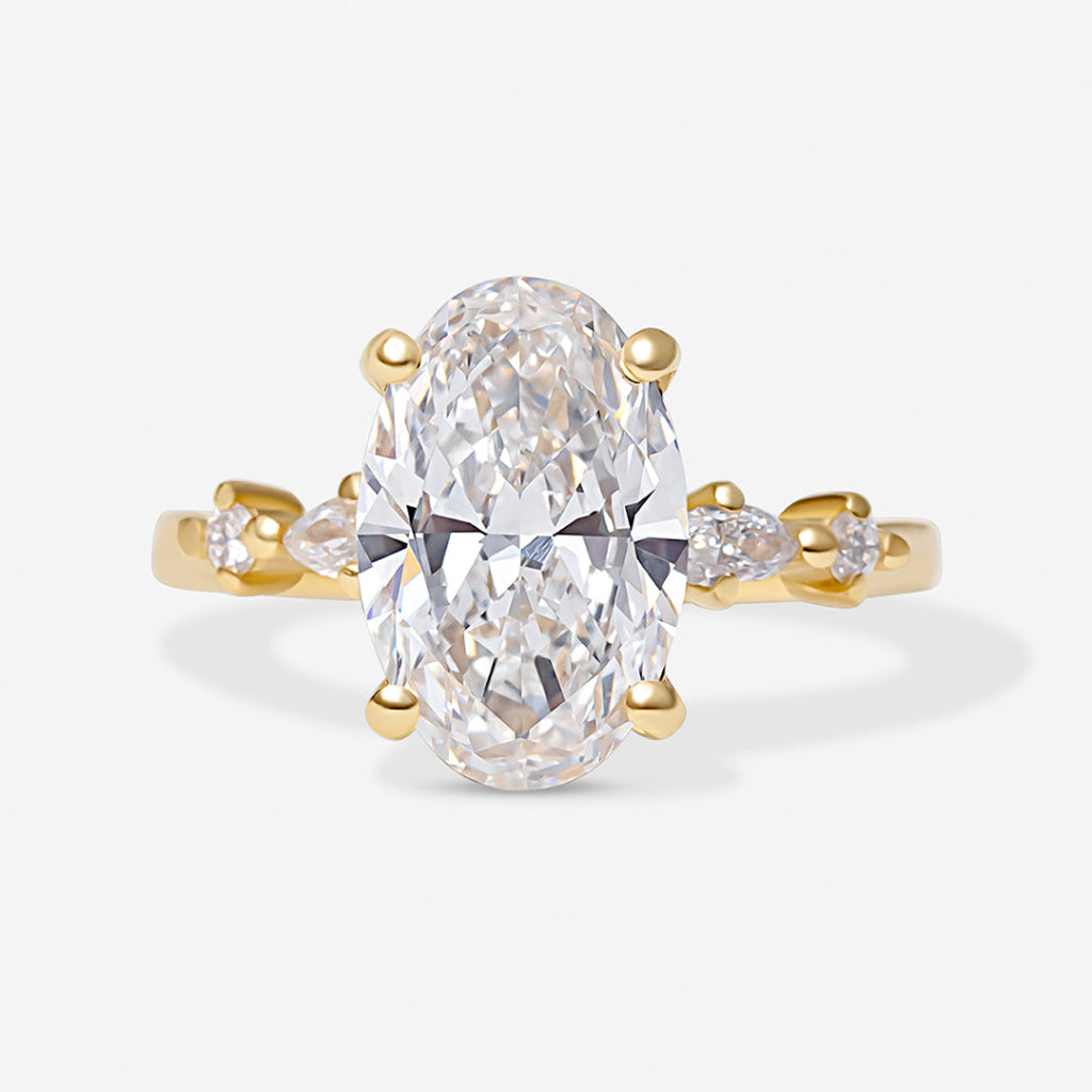An oval-cut diamond engagement ring in yellow gold, featuring a large central oval diamond and smaller marquise-cut diamonds on the shoulders, set against a light gray background.
