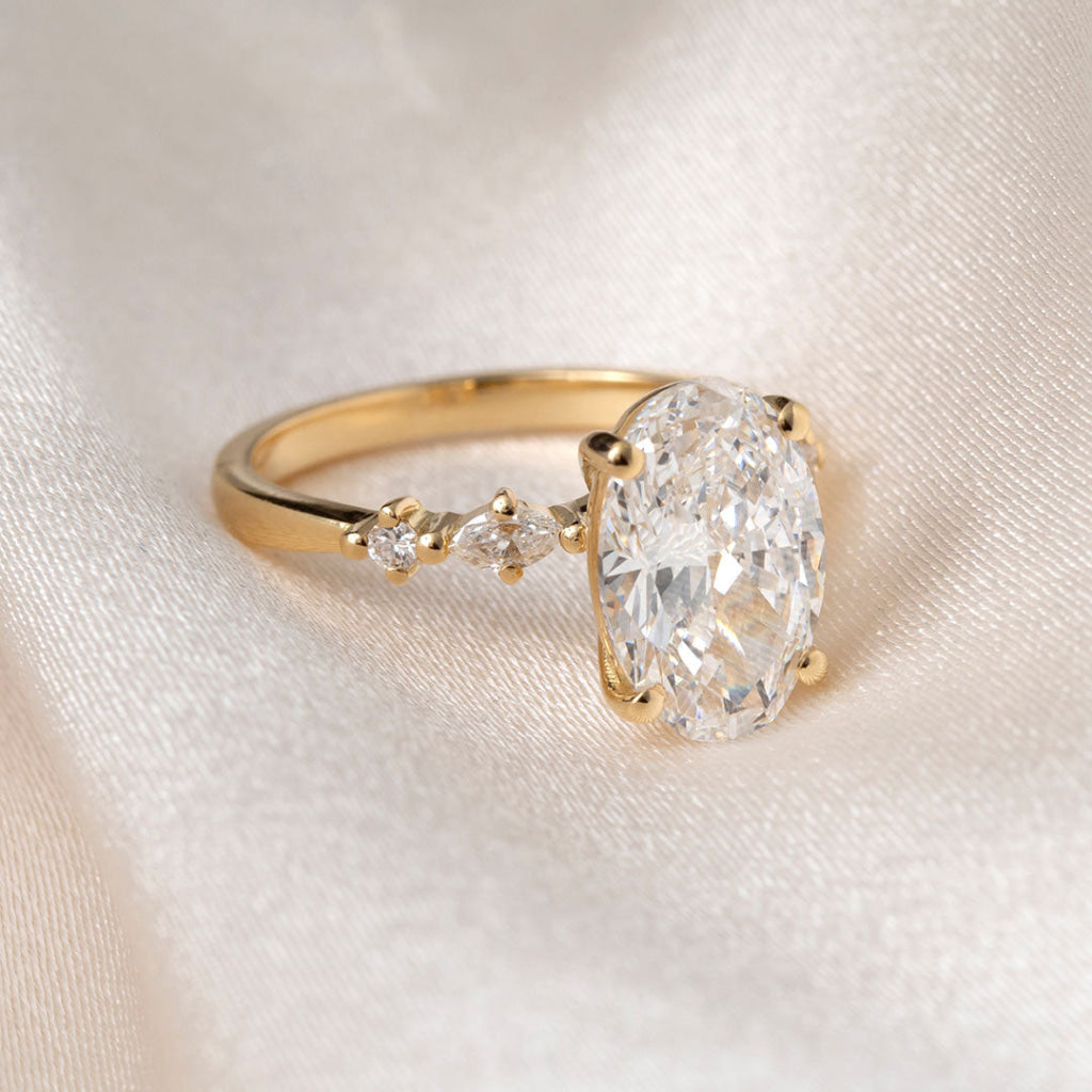An oval diamond engagement ring with a yellow gold band, resting on draped cream-colored satin fabric.
